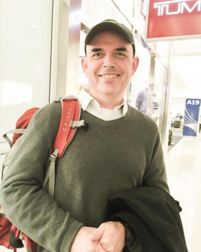 A man wearing a green sweater, white collared shirt, and baseball cap smiles while holding a jacket and wearing a red backpack.