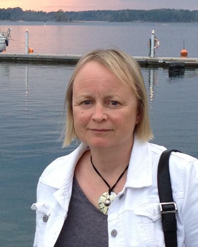 A woman with blonde hair and a white jacket stands in front of a body of water.