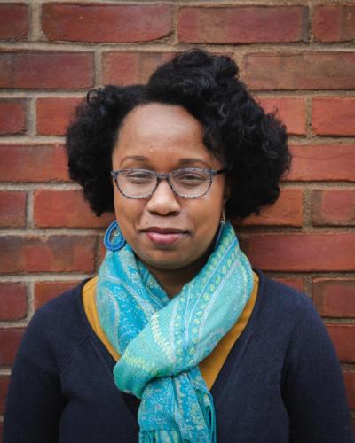 A woman with short, curly hair and glasses stands in front of a brick wall.