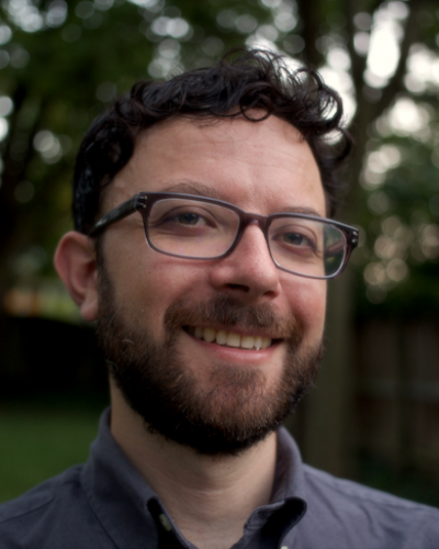 A smiling man with dark hair and glasses stands in front of a blurred background of trees and grass.