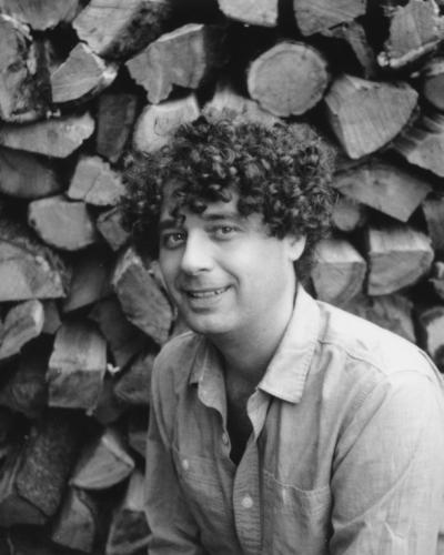 A man with curly hair smiles while leaning in front of a large pile of cut wood.